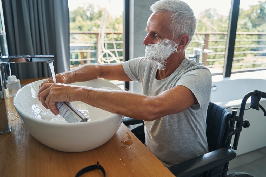 man in wheelchair shaving his face at sink
